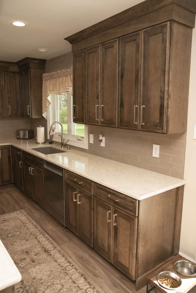A modern kitchen with brown wooden cabinets, a white marble countertop, stainless steel sink, dishwasher, paper towel holder, and a window with beige curtains. Two pet bowls are on the floor near the cabinet.