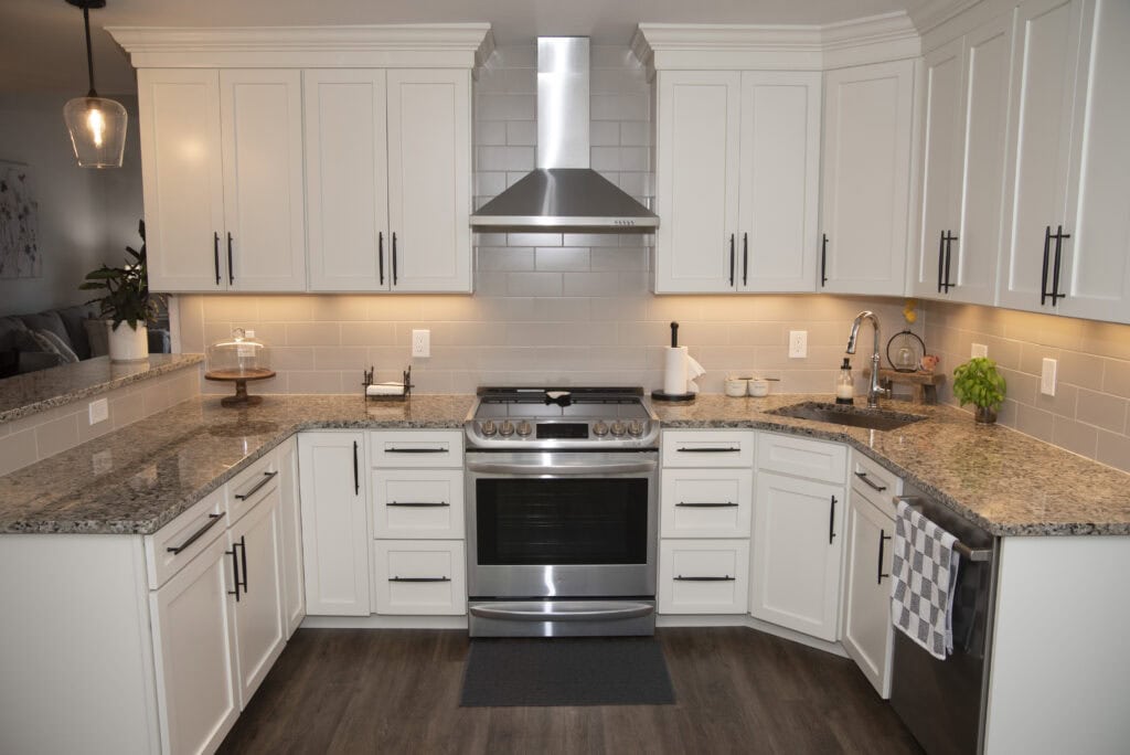 Modern kitchen with white cabinets, stainless steel appliances, granite countertops, subway tile backsplash, under-cabinet lighting, and wood flooring. A stove is centered beneath a range hood, with various kitchen items on the counters.