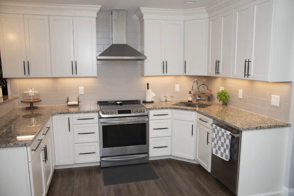 Modern kitchen with white cabinets, stainless steel appliances, granite countertops, a stove with a hood, under-cabinet lighting, a sink, and checkered dish towel hanging on a drawer. Dark wood floors.