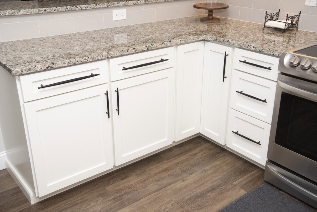 A modern kitchen corner with white cabinets and black handles, granite countertops, a stainless steel oven, and wood-style flooring. A cake stand and napkin holder sit on the counter.