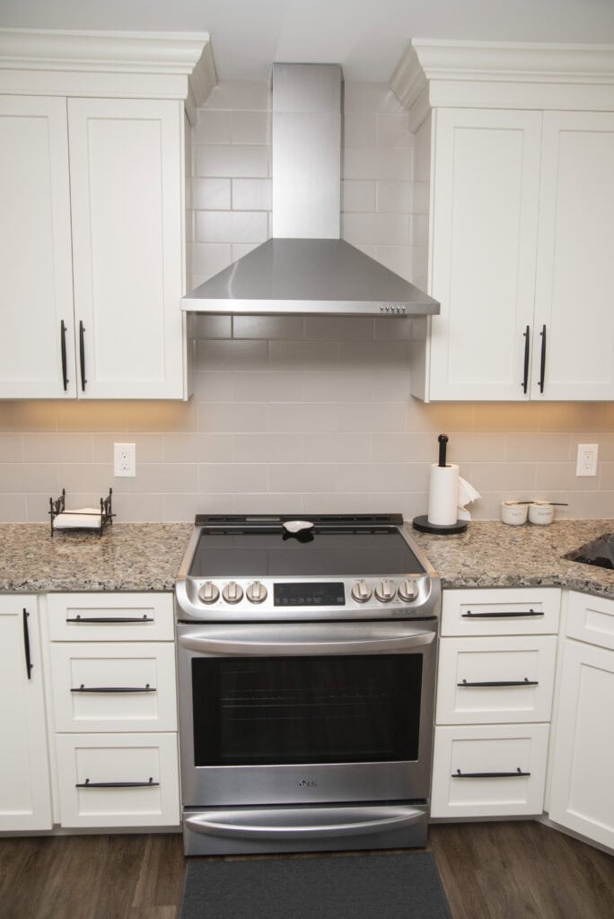 Modern kitchen with white cabinets, granite countertop, and a stainless steel oven and range hood centered against a light gray tile backsplash. A paper towel holder and small containers sit on the right countertop.