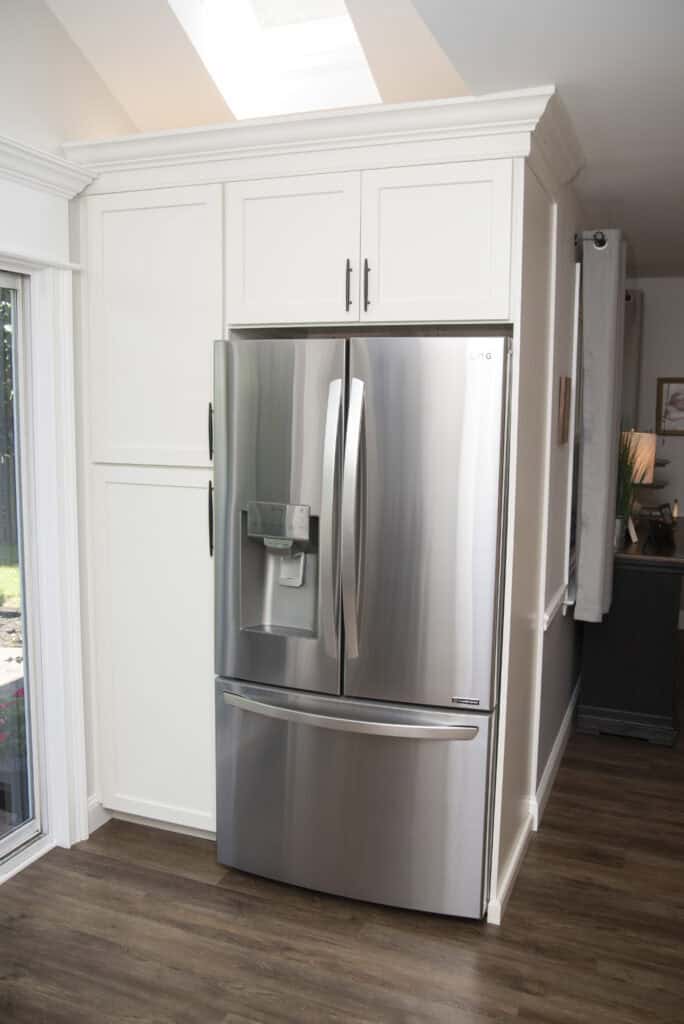 A modern stainless steel French door refrigerator with a water and ice dispenser is built into white kitchen cabinetry, set on wood flooring near a bright sliding glass door.