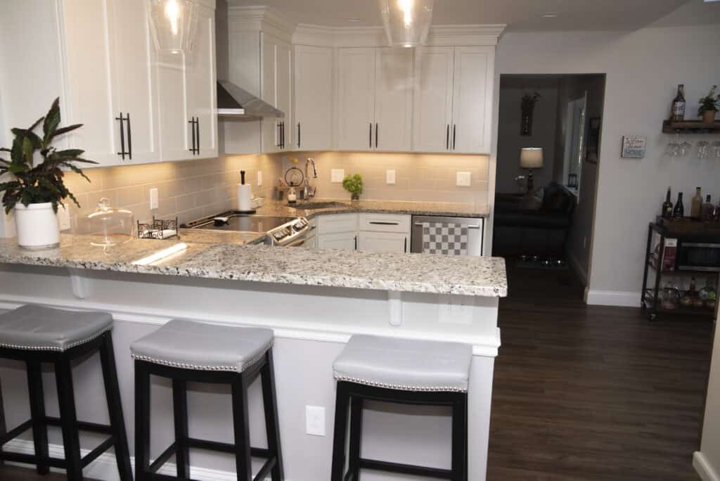 Modern kitchen with white cabinets, granite countertops, and three gray cushioned bar stools. A plant, cake stand, and kitchen essentials are on the counter. In the background, there’s a living area with a lamp and a small bar cart.