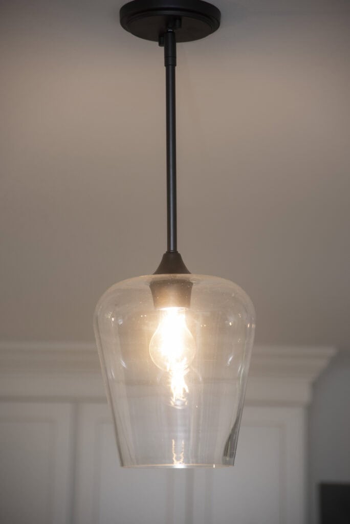 A close-up of a lit pendant light with a clear glass shade hanging from the ceiling, illuminating a room with white cabinetry in the background.