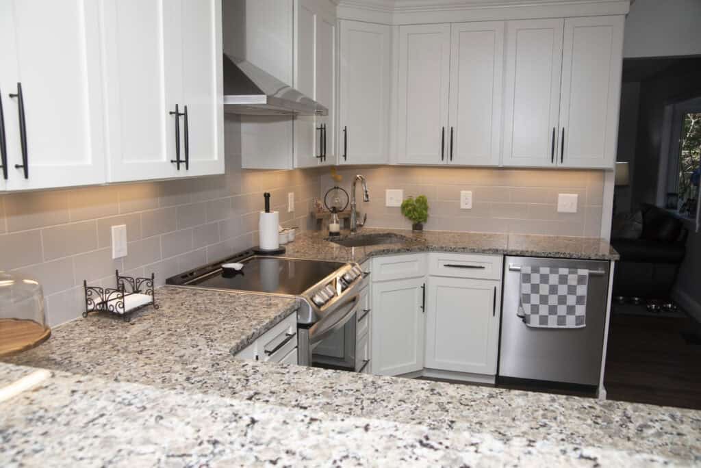 Modern kitchen with white cabinets, granite countertops, stainless steel stove and dishwasher, under-cabinet lighting, and a checkered towel hanging on the dishwasher. A plant and utensils are on the counter.