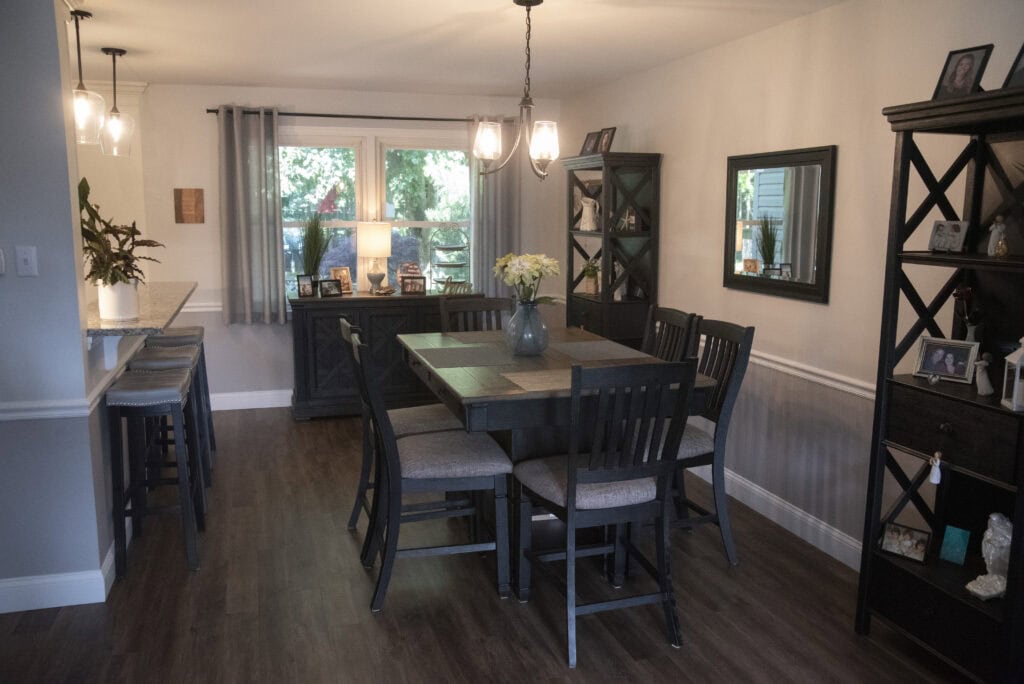 A modern dining room with a wooden table, six chairs, dark wood shelves, framed photos, a vase of flowers, and large windows letting in natural light. The space has pendant lights and hardwood flooring.