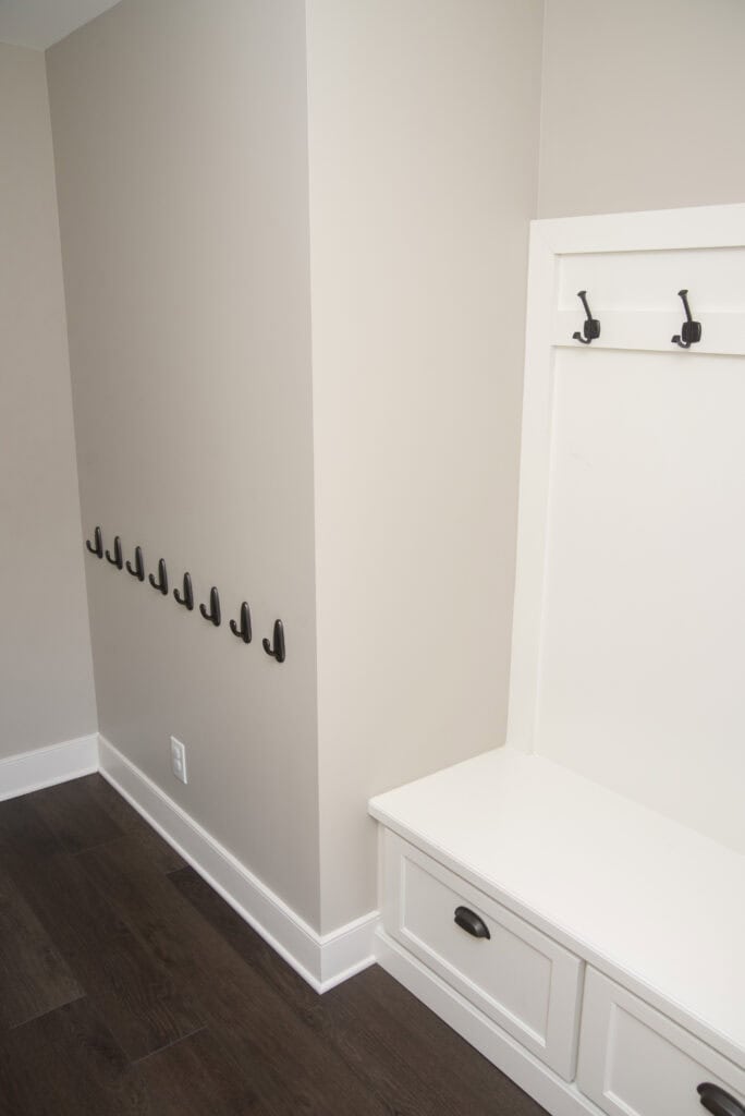 A mudroom with a white built-in bench, two drawers, two wall hooks above, and a row of black hooks along a beige wall, featuring dark wood flooring and white trim.