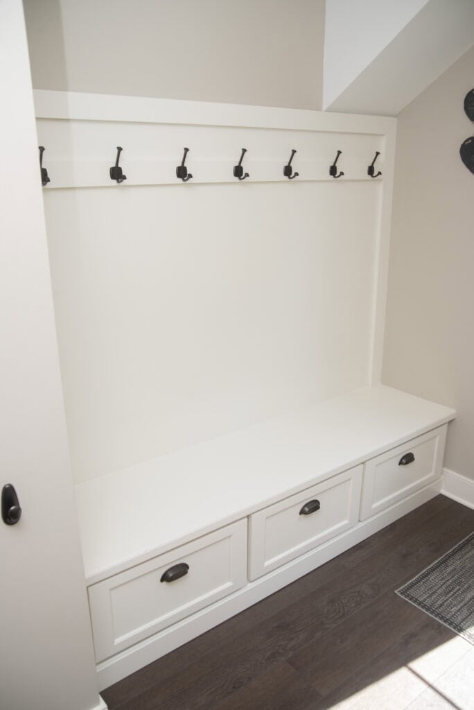 A white mudroom bench with three drawers below and a row of black hooks above, set against a beige wall with dark wood flooring.