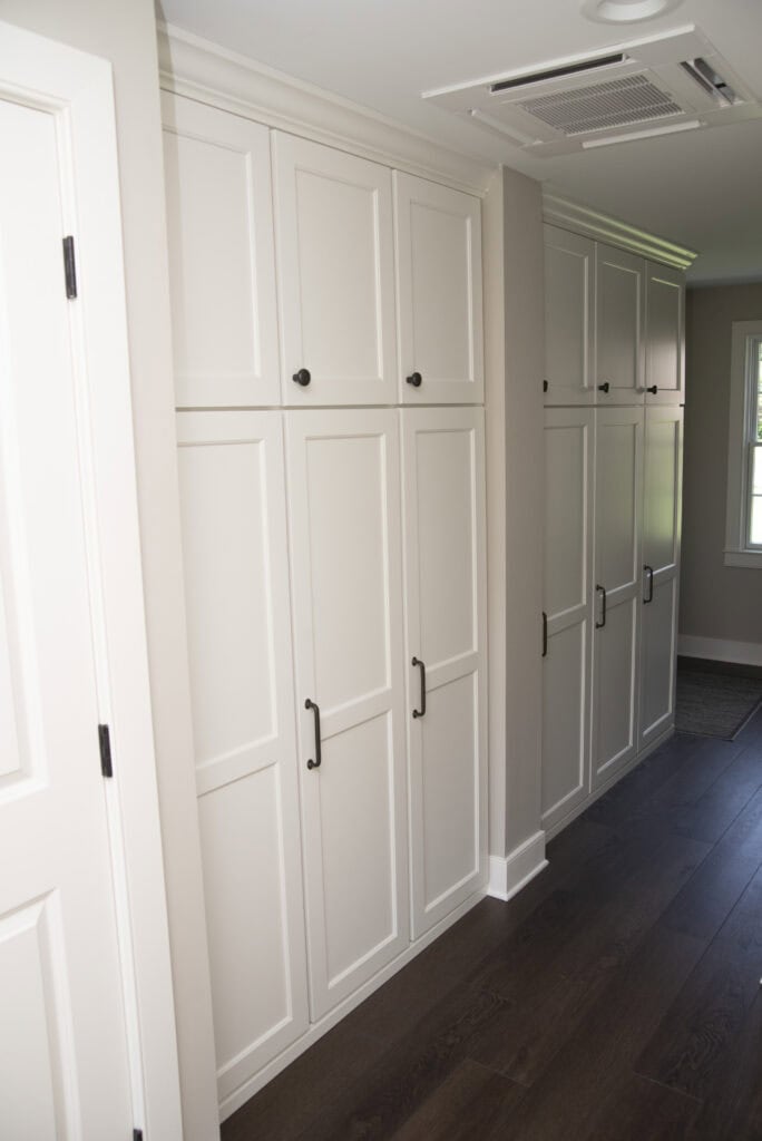 A hallway with white built-in cabinets featuring black handles and knobs, white walls, dark wood flooring, and a window letting in natural light at the end of the corridor.