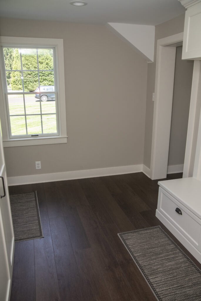 A room with dark wood flooring, beige walls, two gray rugs, a window showing green grass and a parked car outside, and a built-in white cabinet with a drawer on the right.