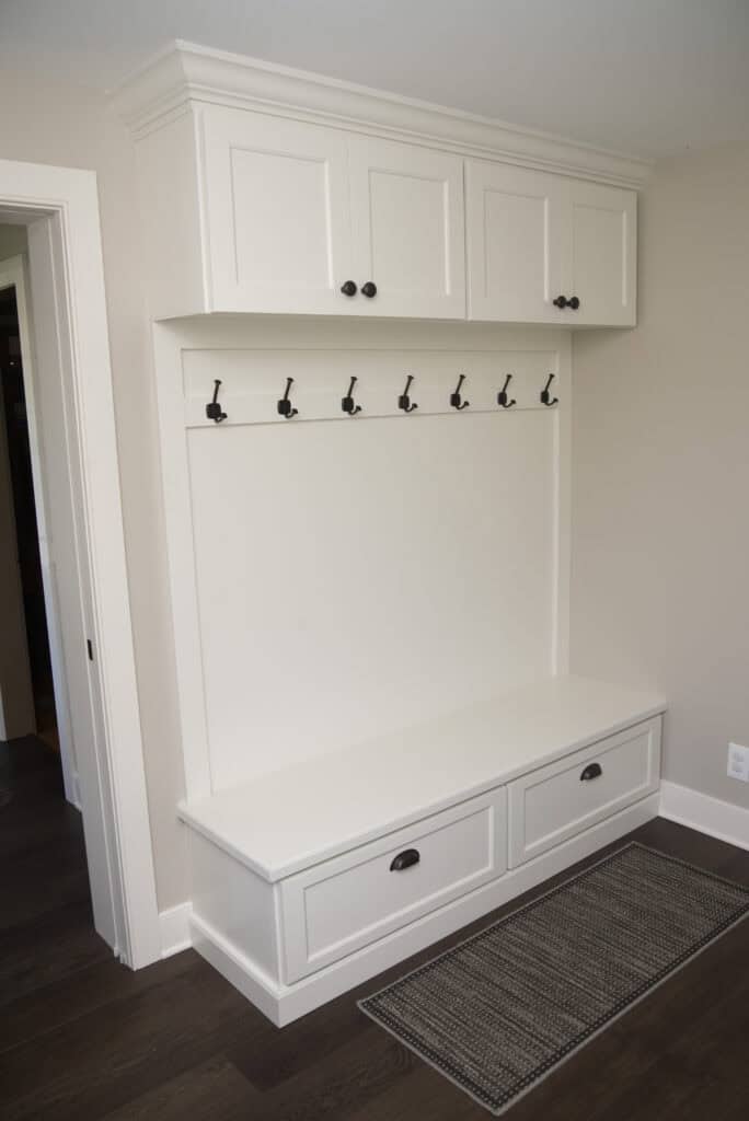 A white mudroom bench with two drawers, a row of six black hooks above, and cabinets overhead. The bench sits against a beige wall with dark wood flooring and a rectangular woven rug in front.