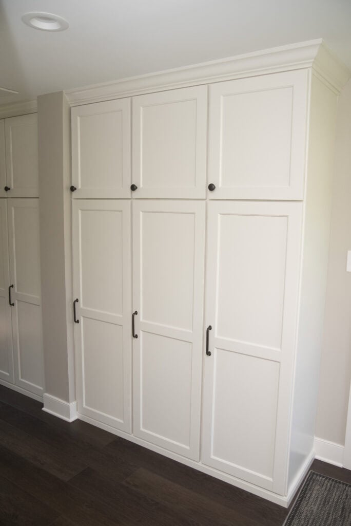 Tall built-in white cabinets with six doors and black handles, set against a light-colored wall on dark wood flooring. The cabinets have a simple, modern design and crown molding on top.