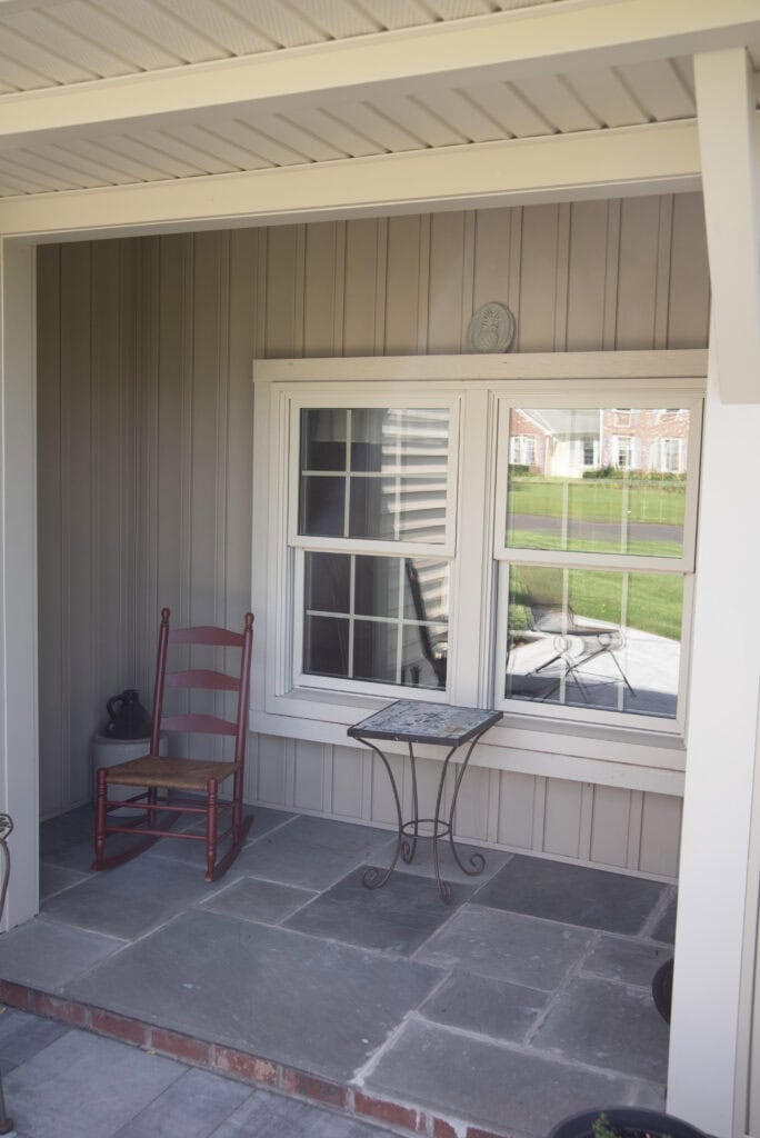 A small porch with a red wooden chair and a metal table in front of a window, set against beige siding and stone flooring. Green grass and houses are visible through the window reflection.