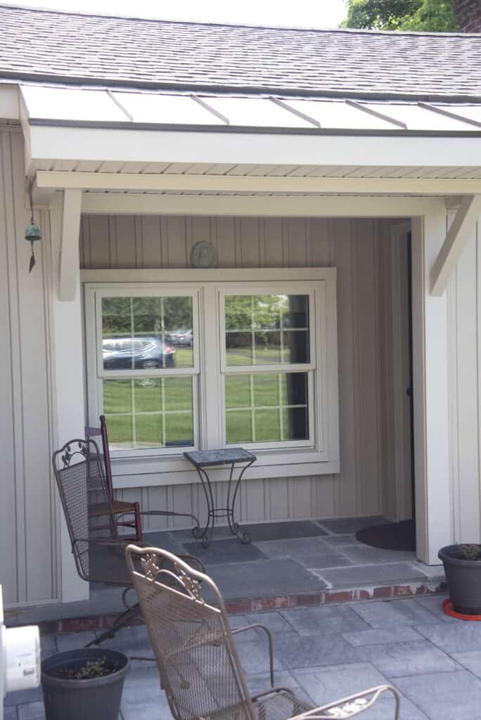 A small covered porch with two metal chairs, a small table between them, and a window facing the porch. The porch has gray stone flooring and potted plants. A grassy lawn and parked cars are visible through the window.