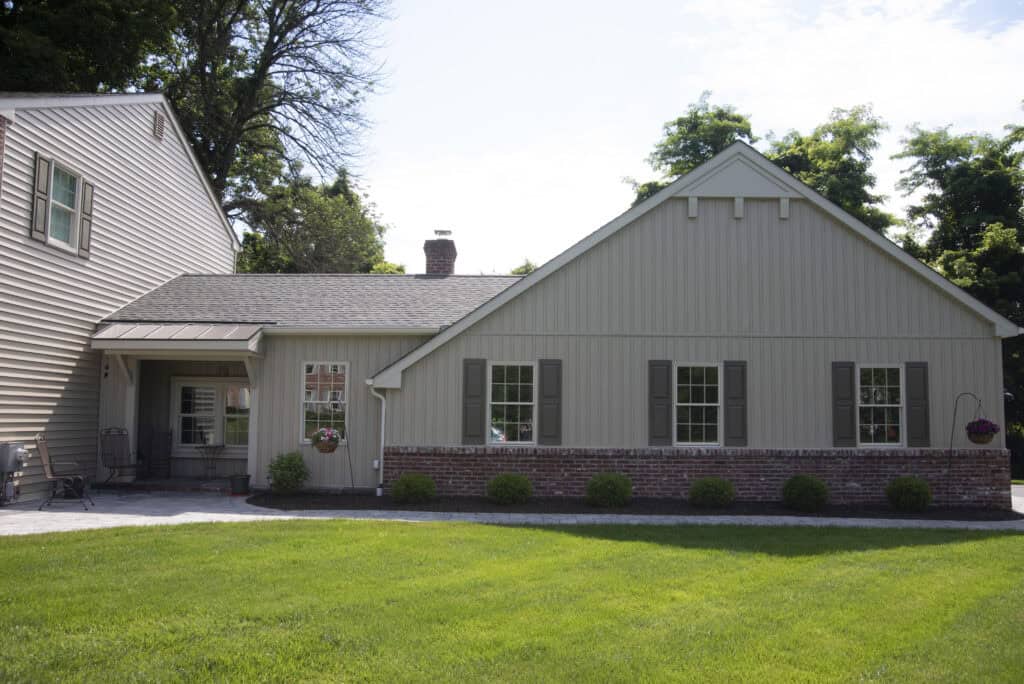 A single-story beige house with brown shutters, a brick lower facade, and a well-manicured green lawn in front. There are a few bushes along the house and trees in the background.