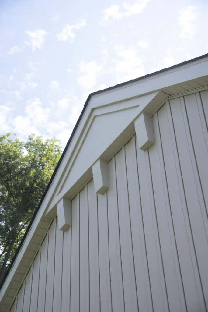 Close-up view of the upper exterior of a modern light beige building with vertical siding and three decorative support beams, set against a partly cloudy sky and trees in the background.