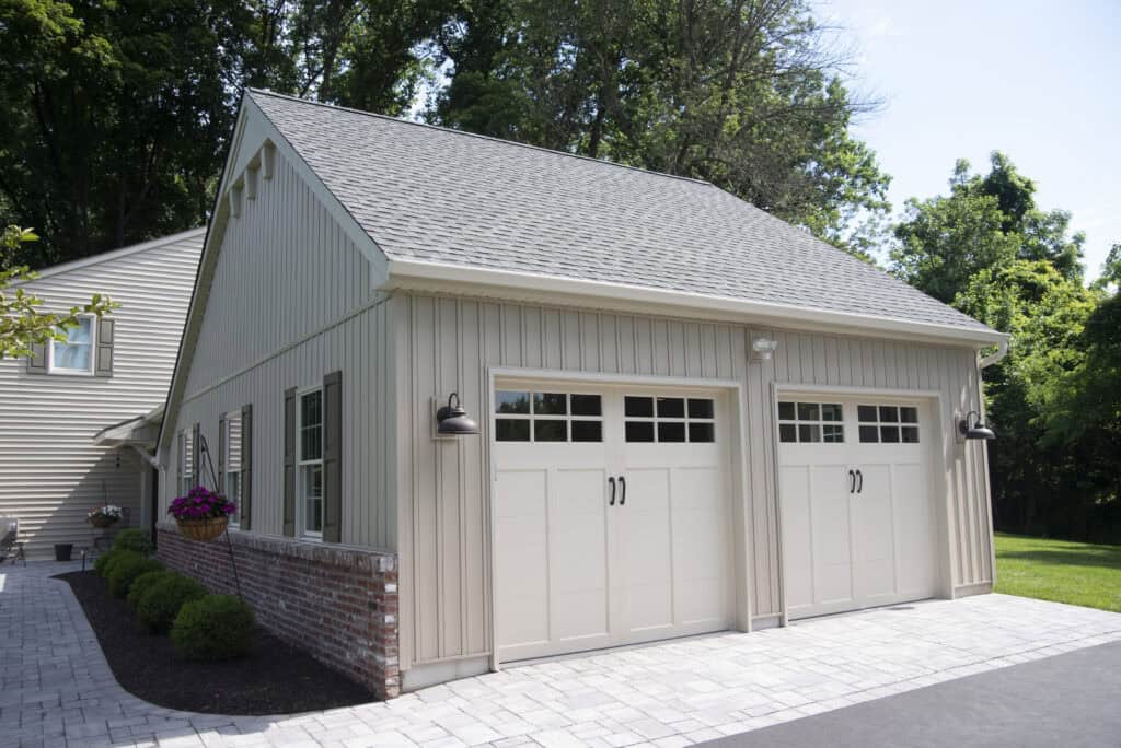A beige detached garage with two white carriage-style doors, black handles, outdoor wall lanterns, and a brick accent base, set beside a driveway and surrounded by green trees and landscaping.