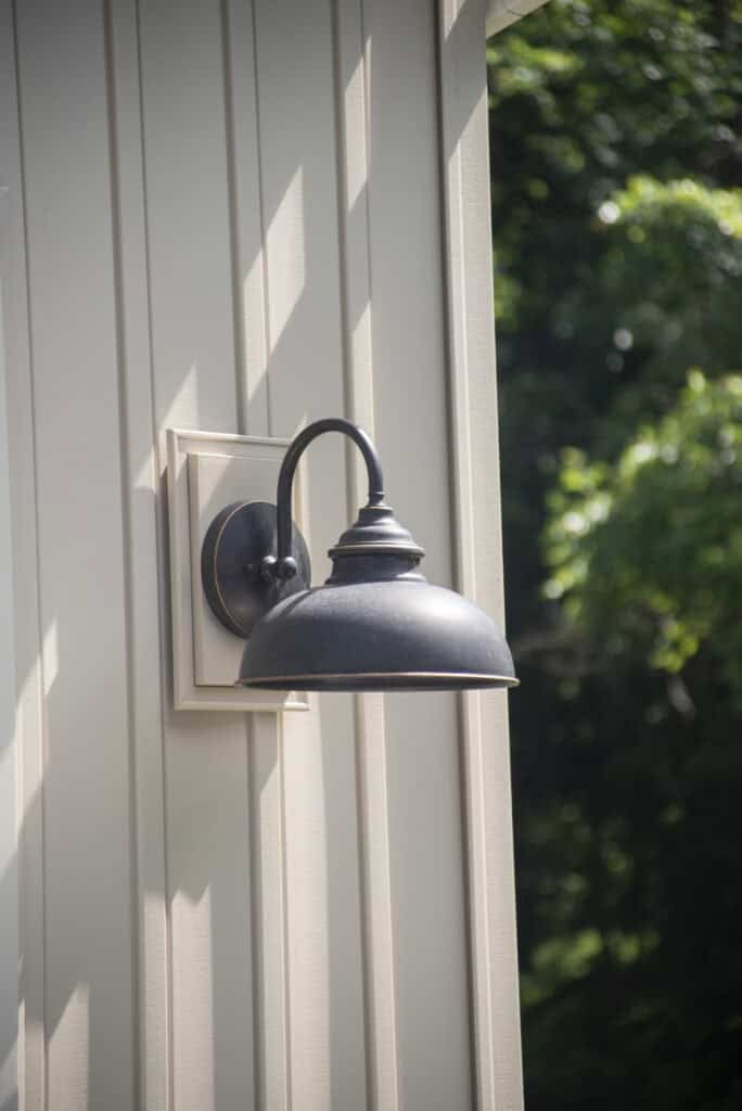 A black outdoor wall lantern with a curved arm is mounted on light-colored vertical siding, with green trees visible in the blurred background.