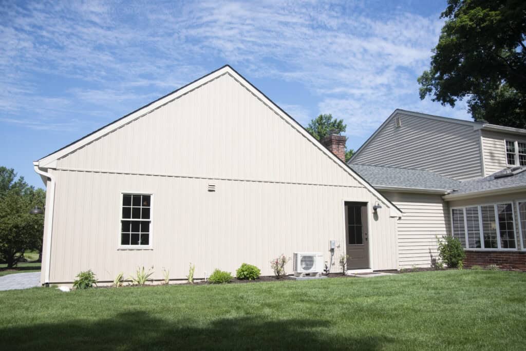A beige house with a sloped roof, one window, a gray door, and an air conditioning unit on a well-kept lawn under a blue sky with scattered clouds.