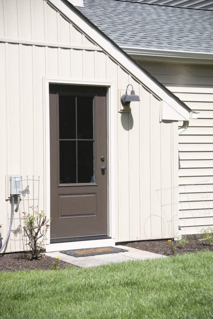 A brown door with window panes is set into beige siding on a house, with a black outdoor light above it, small plants by the entrance, and a doormat on a concrete step leading to a grassy yard.