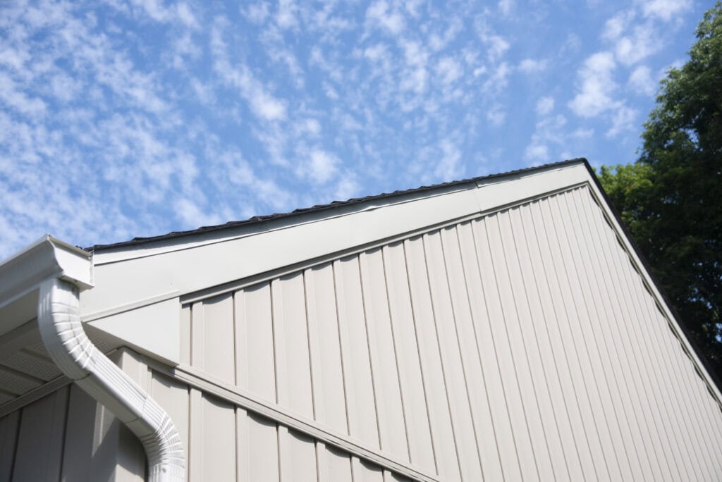 A close-up view of a beige buildings sloped roof and siding, with a white rain gutter. The sky above is blue with scattered white clouds and a tree is visible on the right.