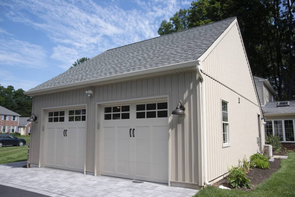 A beige detached two-car garage with white doors and black handles, surrounded by a paved driveway, green grass, small plants, and trees under a blue sky with scattered clouds.