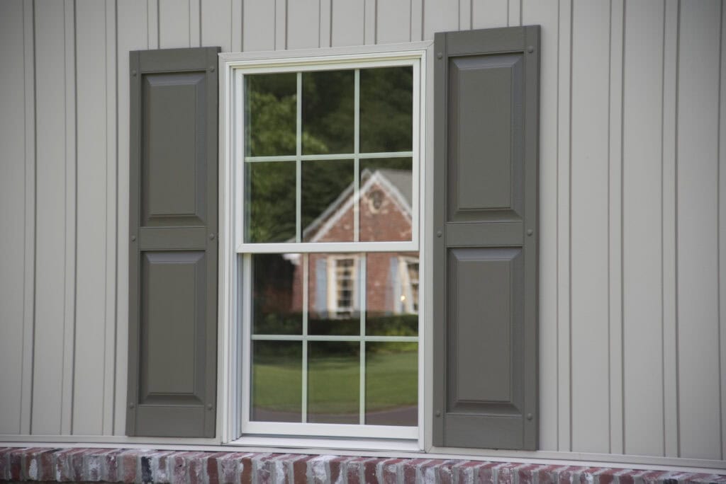 A close-up of a window with gray shutters on a beige house. The window reflects a brick house with white trim, green lawn, and trees in the background. The exterior wall features vertical siding and a brick ledge below the window.