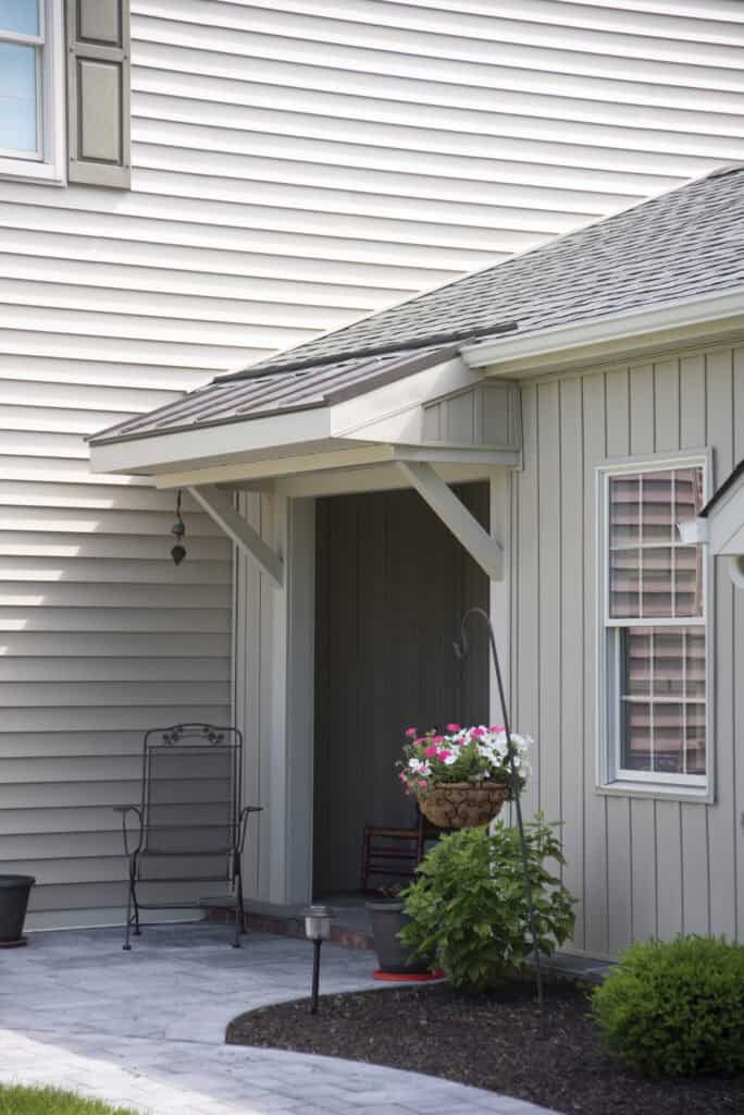 A small covered porch at a house entrance features a black metal chair and a hanging basket of flowers. The light-colored siding and windows enhance the clean, welcoming look, with a garden and walkway in the foreground.