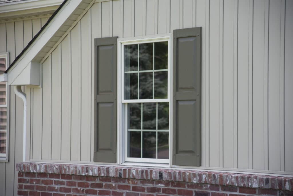 A beige house exterior with vertical siding, a white-framed window with divided panes, olive green shutters on each side, and a red brick foundation. Trees are reflected in the window glass.