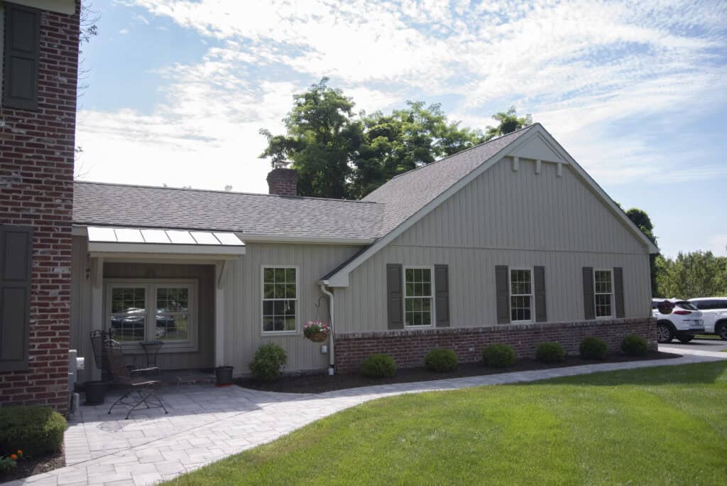 A single-story beige house with brick trim, multiple windows with brown shutters, and a small patio area with chairs. The house is surrounded by green grass, landscaping, and a few trees under a partly cloudy sky.