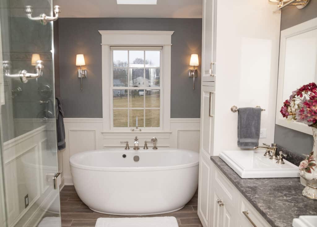 Modern bathroom with a white freestanding bathtub under a window, gray accent wall, wall sconces, marble countertop, white cabinets, and a bouquet of flowers by the sink.