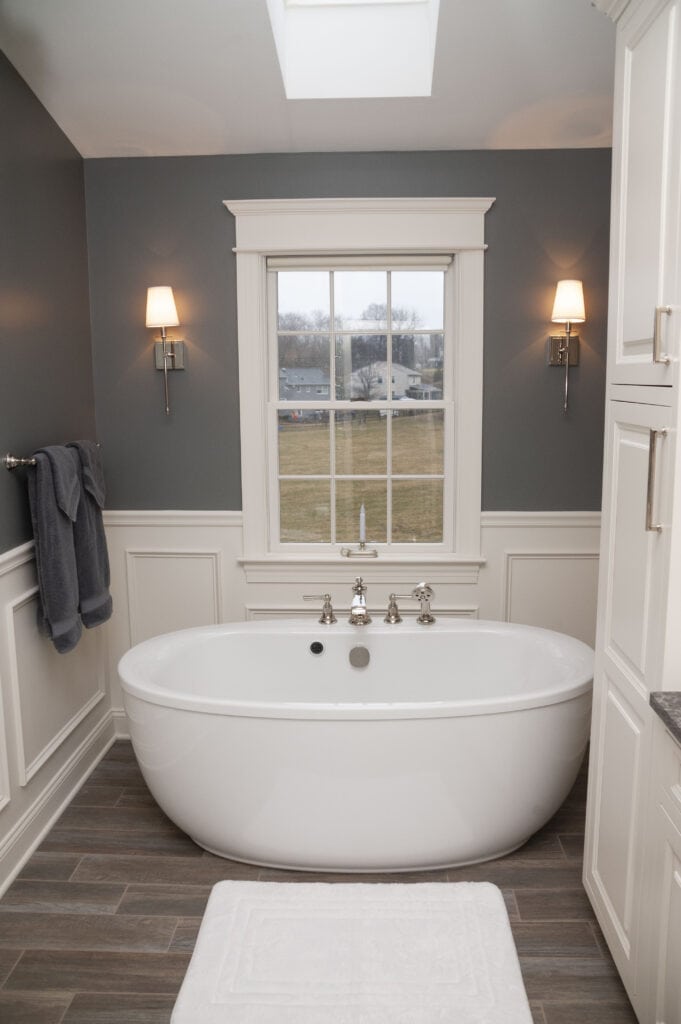 A modern bathroom with a white freestanding tub below a window, gray walls, two wall sconces, folded towels on a rack, wood-look tile flooring, and a white bath mat.