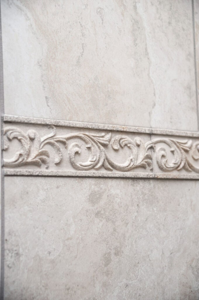 A decorative beige wall tile with a horizontal raised floral and vine pattern running across the center, bordered by smooth tiles on the top and bottom.