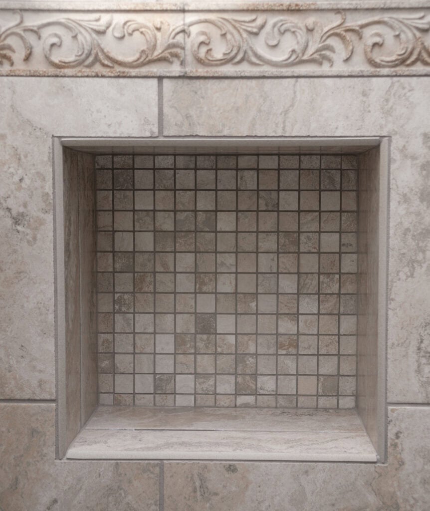 A recessed shower niche with small square beige tiles, surrounded by larger stone tiles. The top edge features a decorative, embossed floral border.