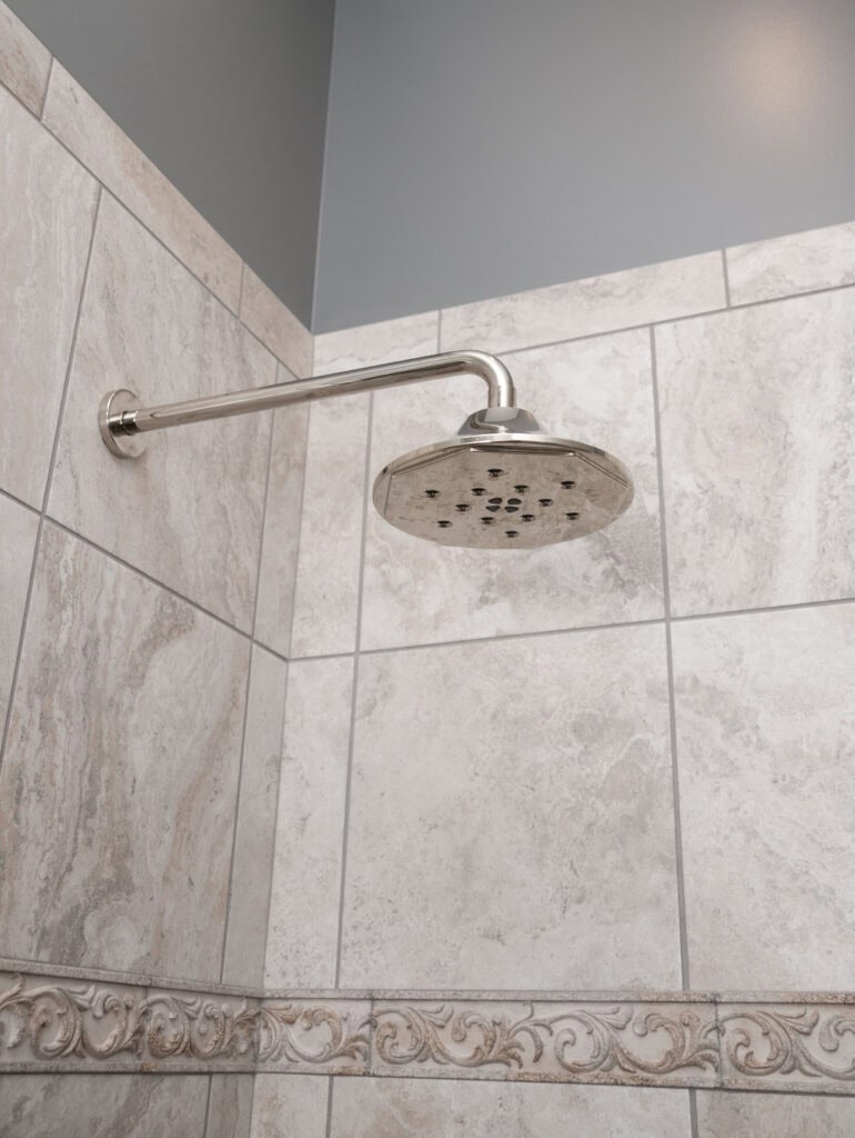 A modern silver showerhead mounted on a tiled bathroom wall with beige and gray marble patterns and decorative trim along the bottom.