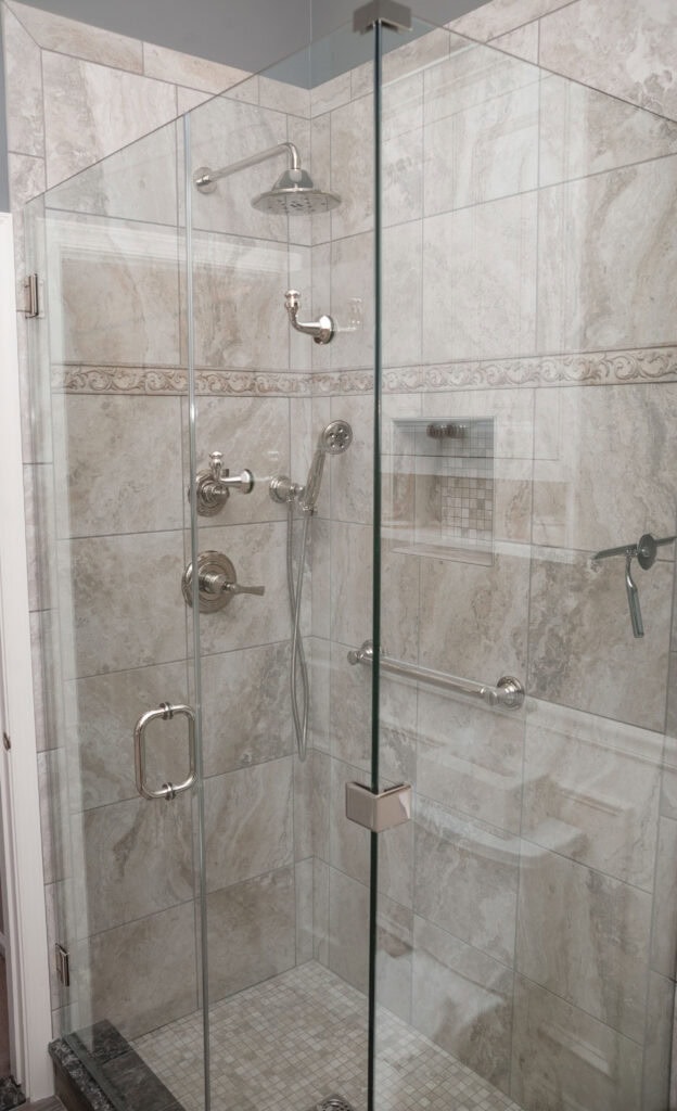 A modern walk-in shower with glass doors, beige and gray tiled walls, a built-in shelf, a rain showerhead, a handheld showerhead, and chrome fixtures.