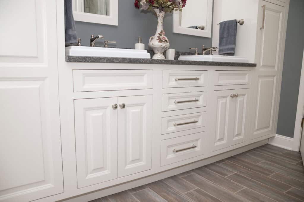 White double-sink bathroom vanity with gray countertop, silver faucets, and cabinet hardware. Blue-gray wall, two mirrors, towels, and a floral vase sit on the counter. The floor has wood-look tiles.