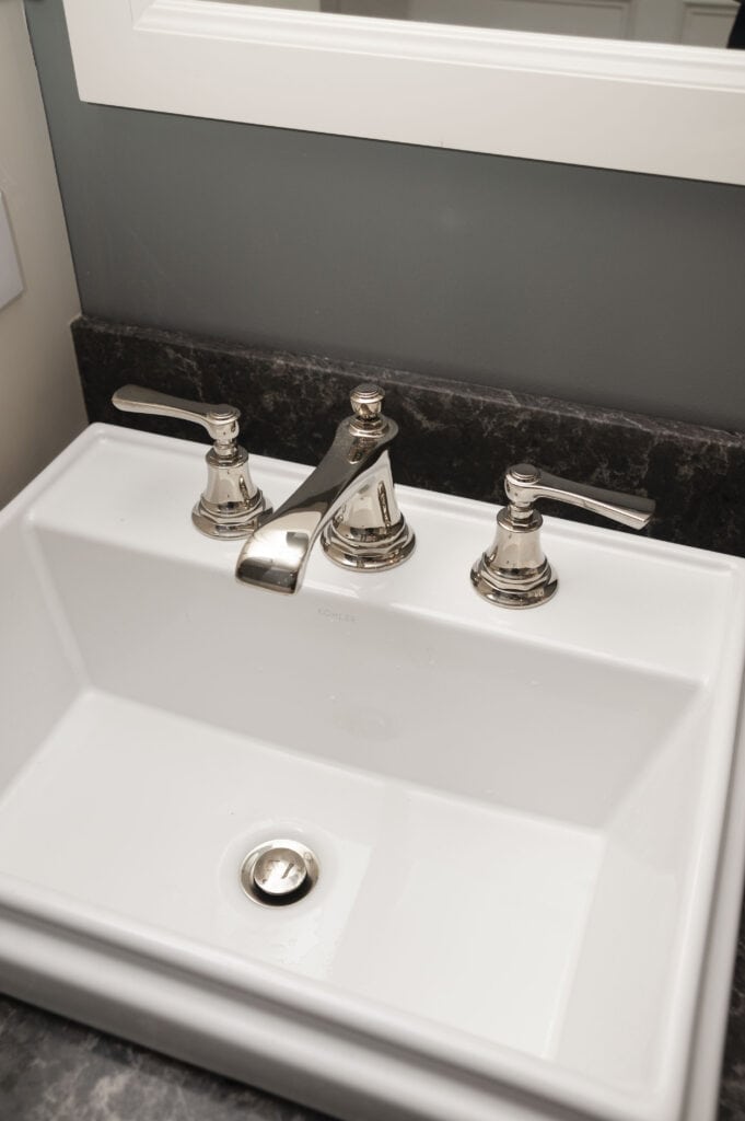 A white rectangular bathroom sink with a silver faucet and two handles, set in a dark marble countertop below a mirror.
