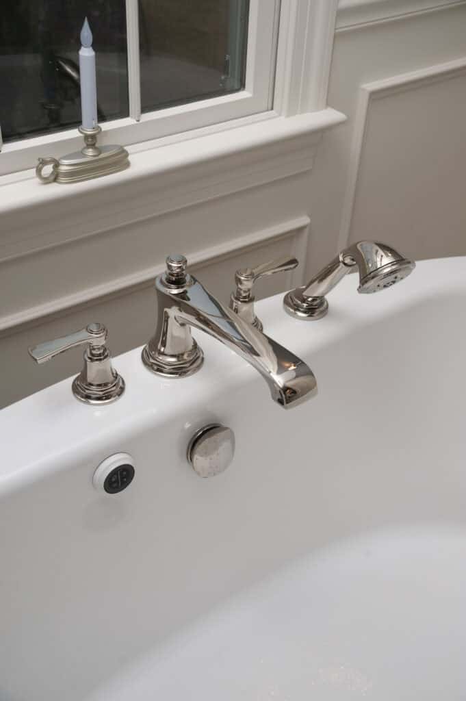 Close-up of a white bathtub with chrome fixtures, including a faucet, two handles, and a handheld shower head, positioned below a window with a white candle on the windowsill.