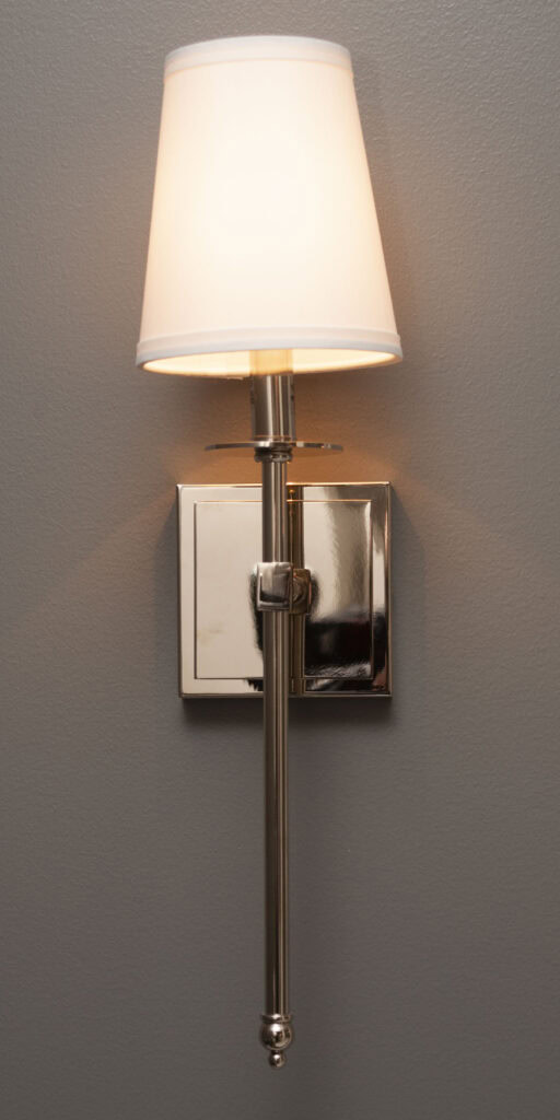 A modern wall-mounted sconce with a polished metal base and a white fabric lampshade, casting warm light against a gray wall.