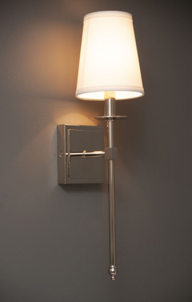 A modern wall-mounted sconce with a metallic base and a white tapered lampshade, glowing warmly against a plain gray wall.
