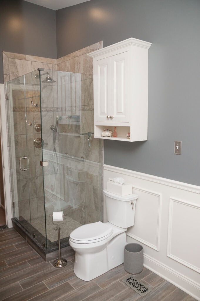 A modern bathroom with gray walls, wood-look tile floor, a glass-enclosed shower with marble tile, a white toilet, wall-mounted cabinet, tissue box, and a small gray trash can.
