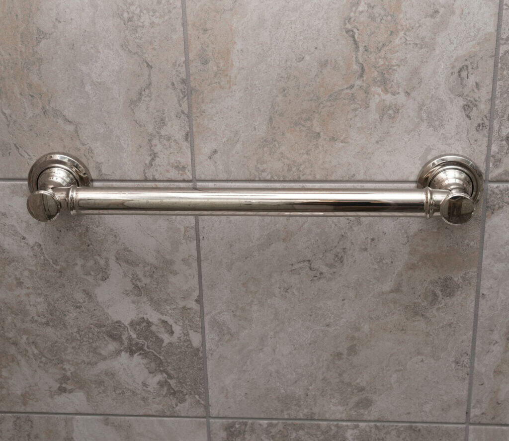 A metal grab bar is mounted horizontally on a tiled bathroom wall, providing support and safety assistance. The tiles have a marbled beige and gray pattern.