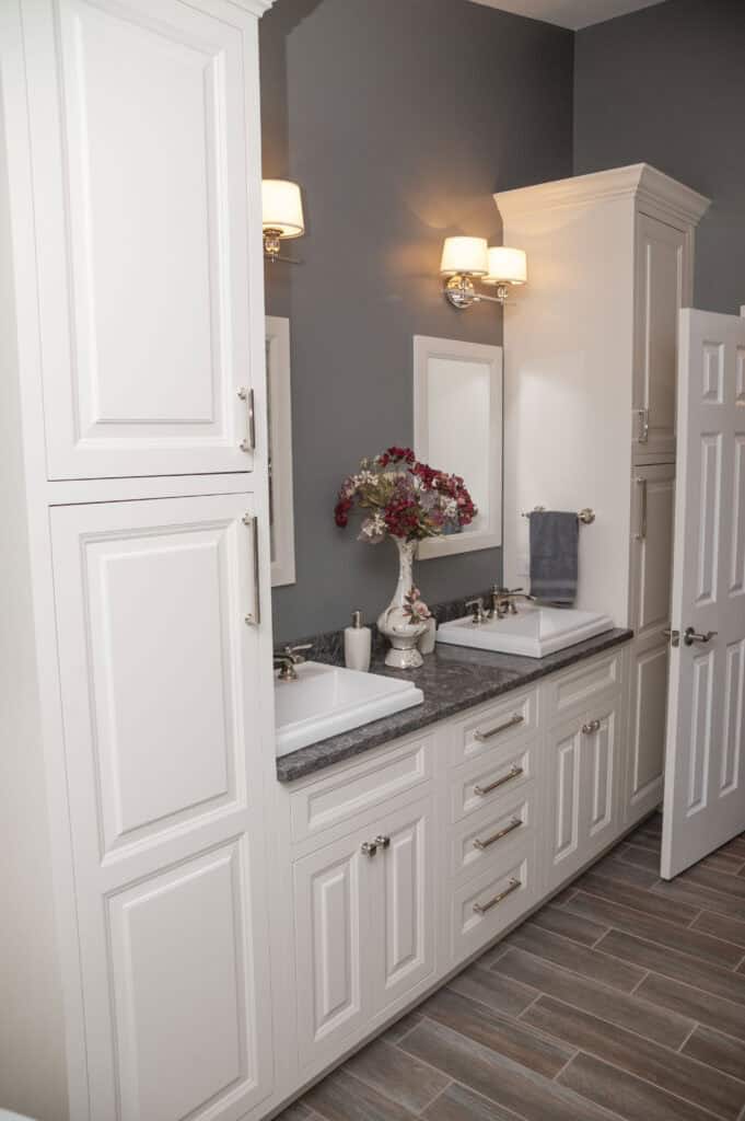 A bathroom with white cabinets, double sinks, gray countertops, wall-mounted lights, a vase of red and white flowers, a towel, and wood-patterned tile flooring.