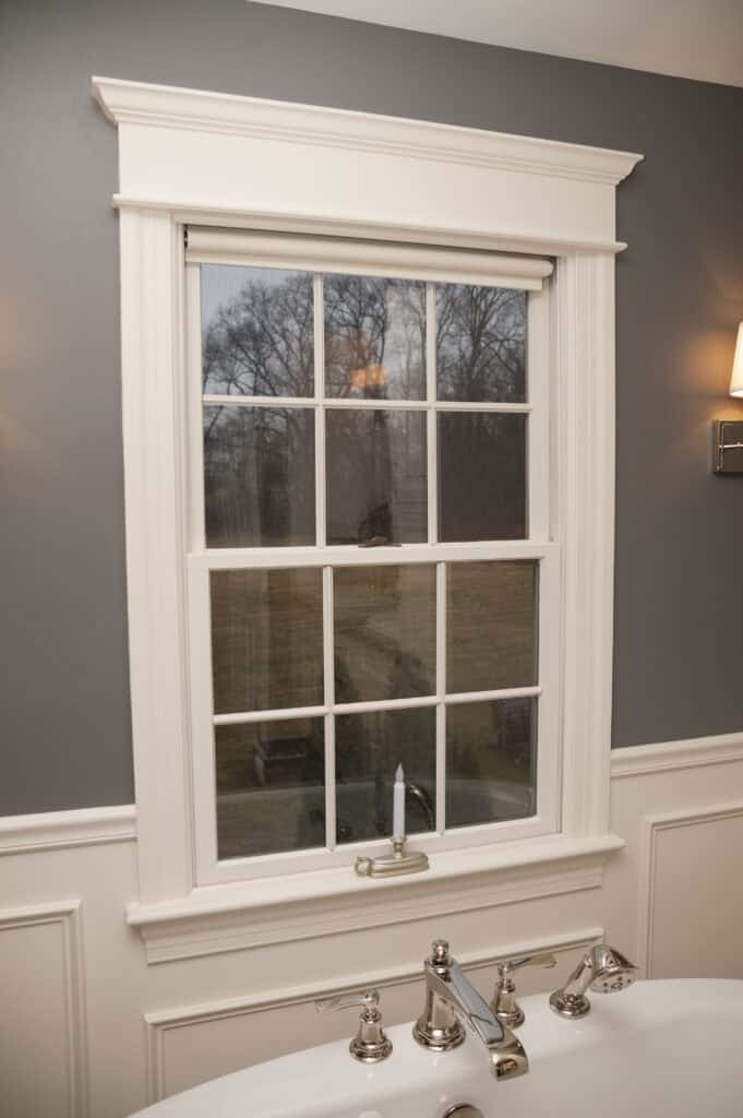 A white-framed window with grid panes is set in a gray and white wall above a bathtub with silver fixtures. Bare trees and a lawn are visible outside through the window.
