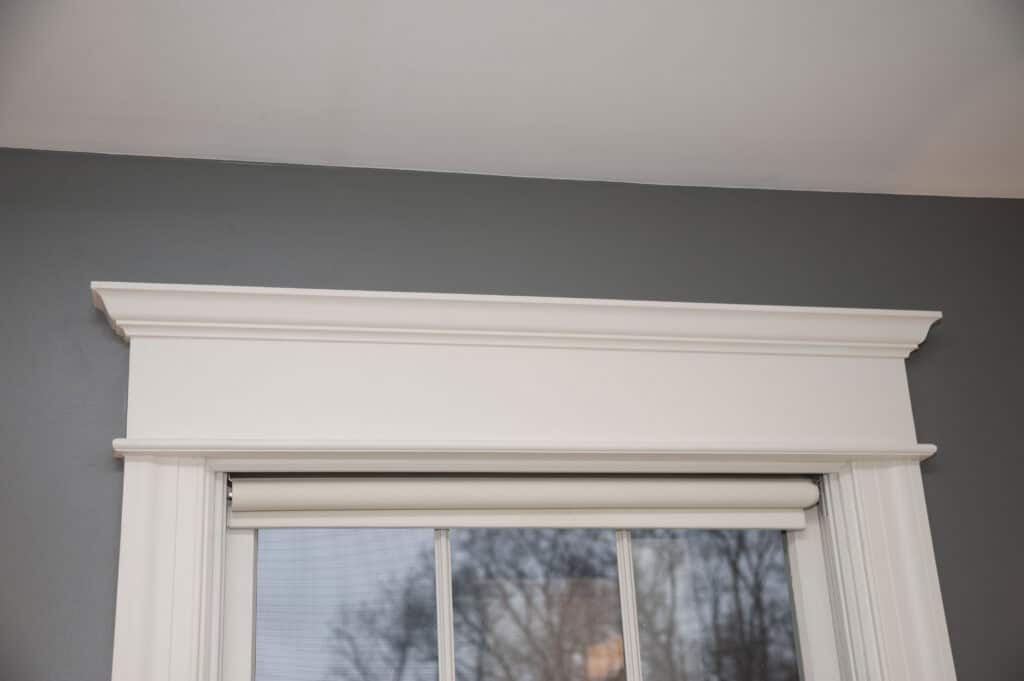 A white, decorative window trim with crown molding frames the top of a window with a rolled-up shade. Outside, bare tree branches are visible against a gray wall and ceiling.