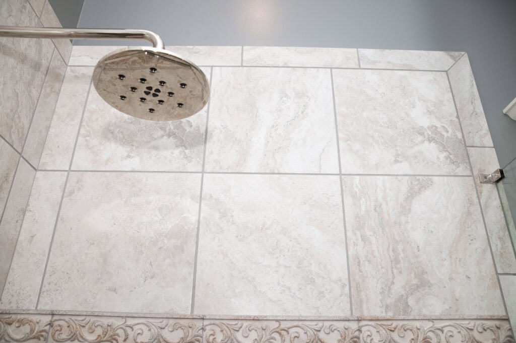A close-up view of a tiled shower wall with a silver showerhead. The tiles are light-colored with subtle patterns, and there is a decorative border along the bottom.