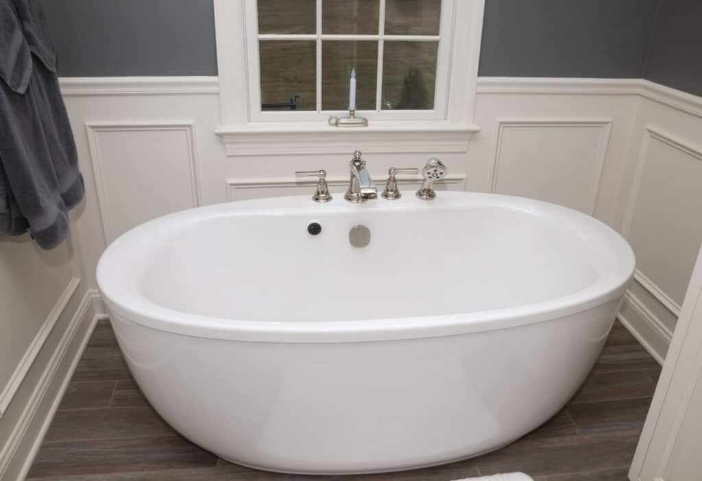 A white freestanding bathtub with silver fixtures sits below a window in a bathroom with white paneled walls, gray upper walls, and brown wood-look tile flooring.