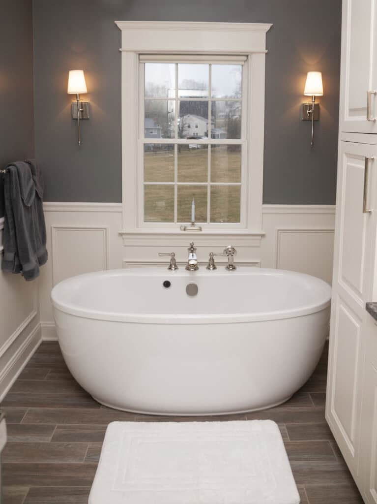 Modern bathroom with a large white freestanding bathtub centered under a window, gray walls with white trim, wall sconces on each side, wood-look tile floor, and a white bath mat in front of the tub.