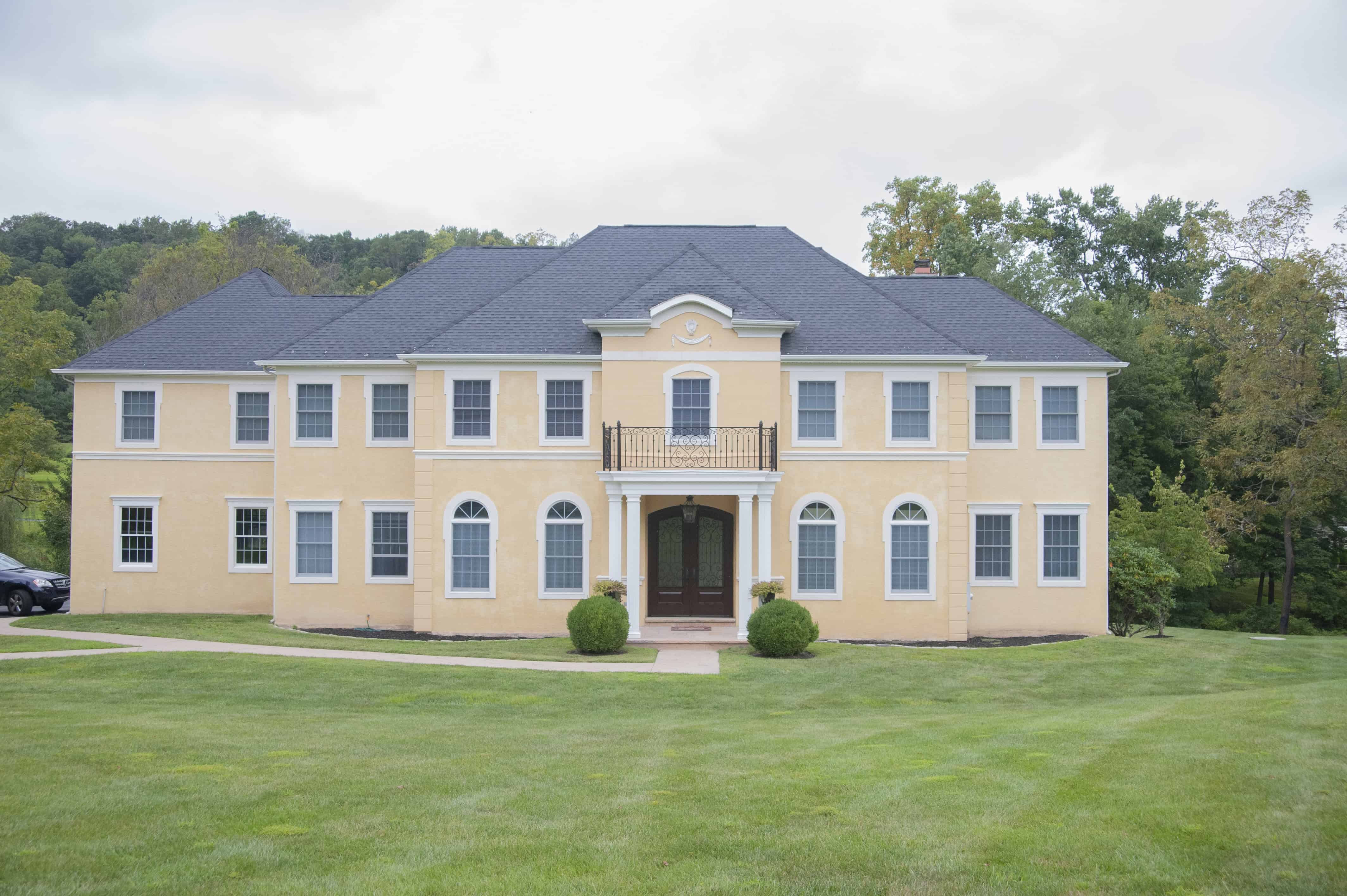 Large, two-story yellow house with white trim, arched windows, and a central entrance with columns, set on a green lawn with trees in the background under a cloudy sky.